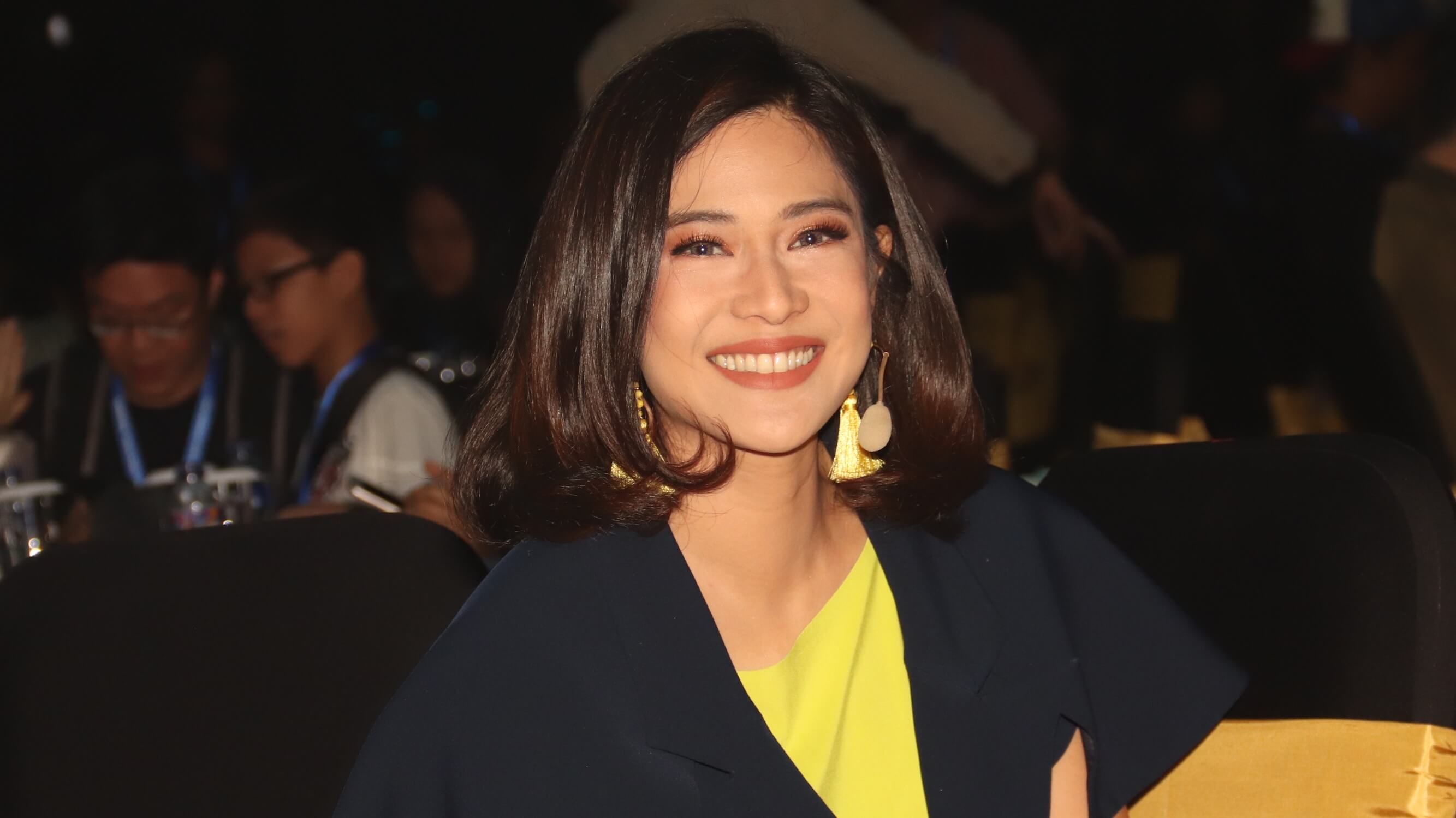 Dian Sastro memakai blouse kuning dengan blazer hitam tersenyum ke kamera